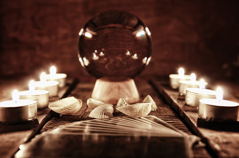 Entorno esotérico, una mesa con varias velas de té encendidas y colocados a ambos lados de la imagen con una bola de cristal en el centro elevada sobre un taco de madera, frente a la bola hay unas conchas de playa blanquecinas y en primer plano unas cartas de tarot repartidas - El Templo de Gala -Tarot
